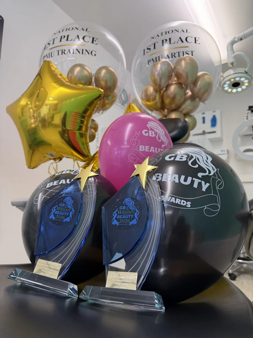 Two blue glass GB Beauty Awards 2026 winner trophies with gold stars on a table, in front of GB Beauty Awards balloons and two clear balloons reading National 1st Place PMU Training and National 1st Place PMU Artist