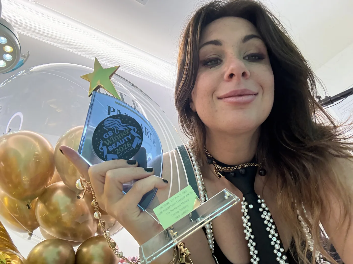 Katy Jobbins smiling and holding up a blue glass GB Beauty Awards winner trophy in front of gold balloons